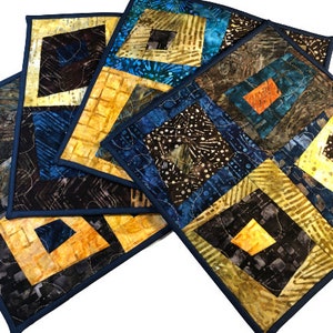 May include: Set of four quilted placemats in a geometric pattern with shades of brown, blue, yellow, and orange. The placemats have a dark blue border.