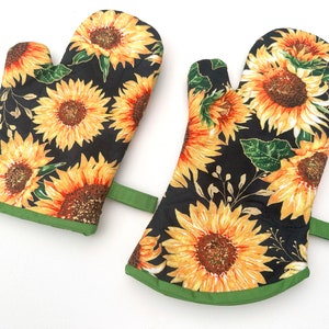 May include: A pair of black oven mitts with a sunflower pattern and green trim. The mitts have a loop on the back for hanging.