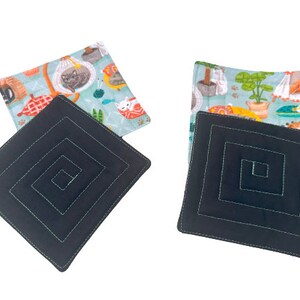 May include: Two black square fabric coasters with a concentric square design. The coasters are topped with fabric squares featuring a light blue background with colorful illustrations of cats and plants.