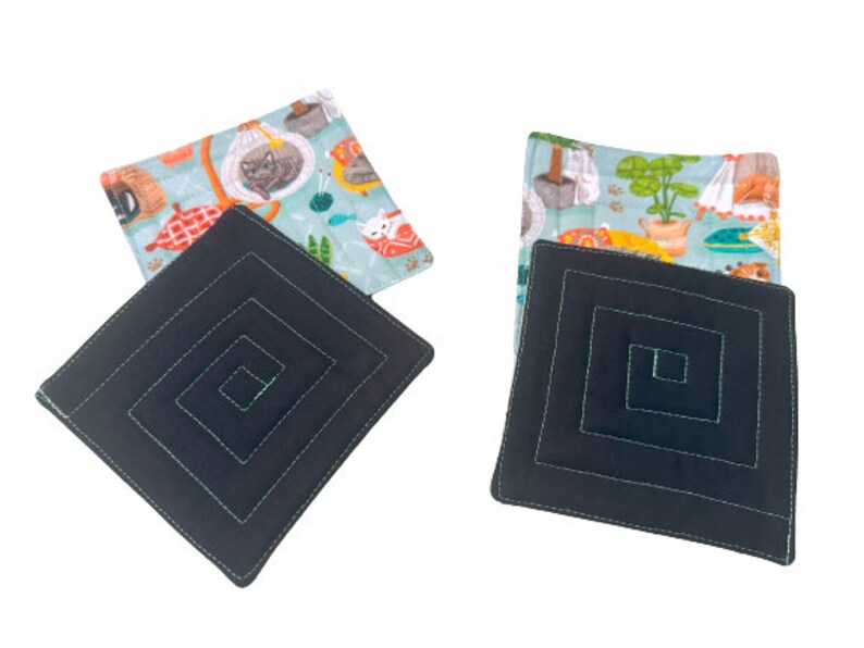 May include: Two black square fabric coasters with a concentric square design. The coasters are topped with fabric squares featuring a light blue background with colorful illustrations of cats and plants.
