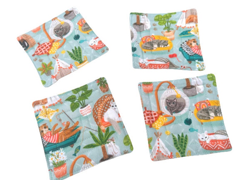 May include: Set of four blue and green fabric coasters with a whimsical cat theme. Each coaster features a different cat design with various colors and patterns. The coasters are perfect for protecting surfaces from heat and spills.
