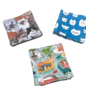 May include: Three square fabric coasters with cat designs. One coaster features a blue background with white cats, another has a green background with cats in various poses, and the third has a brown background with a collage of cat faces.