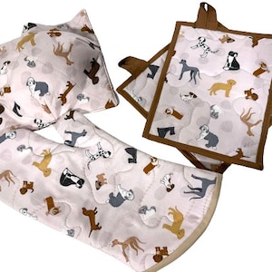 May include: Set of four kitchen accessories featuring a pink quilted fabric with a pattern of various dog breeds. The set includes a pot holder, two hot pads, and a trivet.