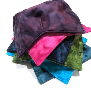 May include: A stack of five colorful quilted pot holders. The top pot holder is a dark purple with a tie-dye pattern. The second pot holder is a bright pink with a tie-dye pattern. The third pot holder is a dark blue with a tie-dye pattern. The fourth pot holder is a green with a tie-dye pattern. The fifth pot holder is a light blue with a tie-dye pattern.