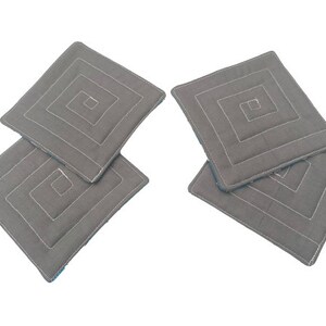 May include: Four gray quilted fabric coasters with a white geometric design. The coasters are stacked on top of each other.