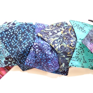 May include: A collection of handmade fabric bowl cozies in various colors and patterns. The cozies feature designs such as floral, cross, and abstract patterns in shades of blue, purple, and teal. The cozies are arranged in a row, showcasing their diverse designs.