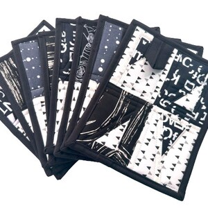 May include: A set of seven square quilted pot holders. Each pot holder features a unique black and white pattern, including geometric shapes, abstract designs, and text. The pot holders have a black border and are arranged in a fan shape.