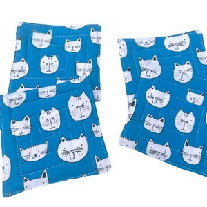 May include: Set of four blue fabric coasters with a white cat face pattern. The coasters are square and have a quilted texture.