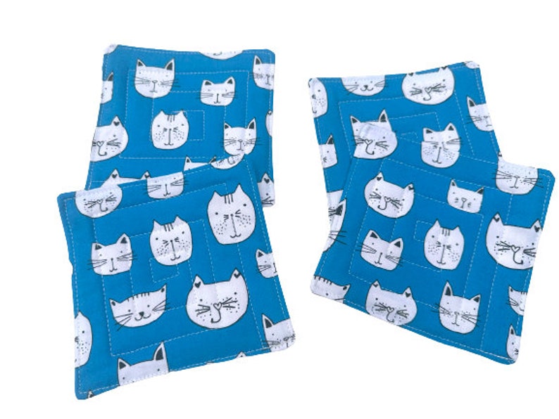 May include: Set of four blue fabric coasters with a white cat face pattern. The coasters are square and have a quilted texture.