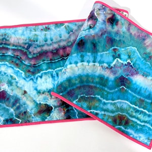 May include: Two tie-dye fabric pieces with a vibrant blue, teal, and purple pattern. The edges are finished with a bright pink border. The fabric has a quilted texture.