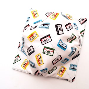 May include: A white fabric bowl with a pattern of colorful cassette tapes. The bowl is folded and appears to be made of a soft, quilted fabric.