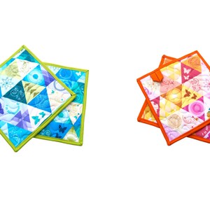 May include: Two square quilted potholders. One set features blue, green, and purple triangles with butterfly and floral patterns, outlined in green. The other set has orange trim and pink, yellow, and white triangles with floral and butterfly patterns.