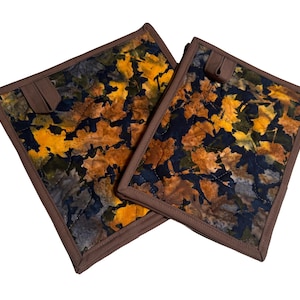May include: Two square quilted pot holders featuring a vibrant autumn leaf pattern in shades of yellow, orange, brown, and dark blue. Each pot holder is finished with a solid brown fabric border and includes a small fabric loop for hanging storage.