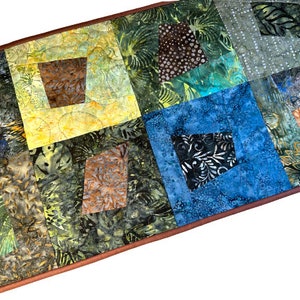 May include: A handmade quilted table runner with a patchwork design. The runner features squares of fabric in shades of green, blue, yellow, and brown. The squares have various patterns, including floral and abstract designs. The edges are finished with a brown border.