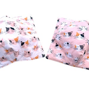 May include: Two pink and white fabric squares with a repeating pattern of colorful cat faces. The fabric squares are folded and appear to be soft and plush.