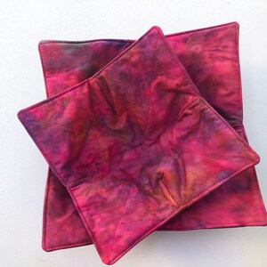 May include: Two quilted fabric bowl cozies in shades of pink, purple, and orange. The cozies are folded and stacked on top of each other.