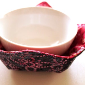 May include: A white ceramic bowl sits inside a quilted fabric bowl cozy. The cozy features a black exterior with a pink floral pattern and a bright pink interior lining with a subtle white pattern, constructed with four folded corner flaps.