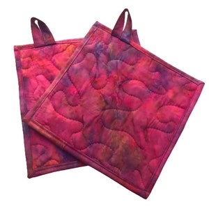 May include: Two quilted hot pads in a pink and purple tie-dye pattern with a loop handle on each.
