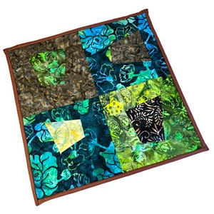 May include: A square, handmade quilt with a brown border. The quilt features a patchwork design with various green, blue, and yellow floral patterns. The quilt is machine quilted with a dark thread.