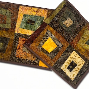May include: Two rectangular quilted table runners featuring a patchwork design of batik fabrics in shades of brown, gold, yellow, and dark green. The runners are finished with a solid dark brown fabric binding along the outer edges.