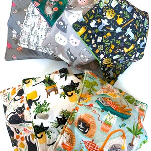 May include: A selection of square fabric bowl cozies featuring cat-themed designs. Patterns include cats amongst flowers, gardening tools, and relaxing scenes. The cozies are in shades of grey, teal, white, and yellow.