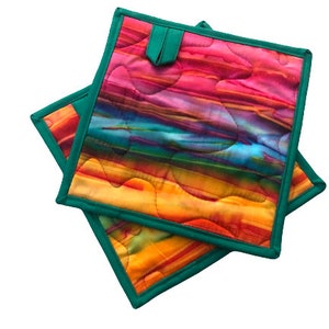 May include: Two colorful quilted pot holders with a teal green border. The pot holders feature a multi-colored abstract design with shades of pink, orange, yellow, blue, and green.