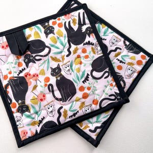 May include: Two square quilted pot holders with a black border. The fabric features a repeating pattern of black cats, floral elements, and Halloween-themed motifs on a light pink background. A black loop is attached.