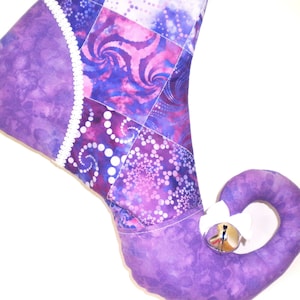 May include: A purple and white Christmas stocking with a whimsical elf shoe design. The stocking features a patchwork pattern with swirling and dotted designs in shades of purple and white. A silver bell hangs from the curled toe.