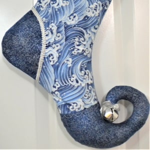 May include: A blue and white wave-patterned Christmas stocking with a denim-like cuff and a silver bell on the curled toe. The stocking is trimmed with silver lace.