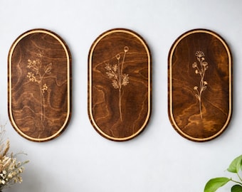 May include: Three oval-shaped wooden wall plaques with a dark brown stained finish and a light-colored border. Each plaque features a delicate, gold-toned line art illustration of a different wildflower, showcasing intricate stems, leaves, and buds.