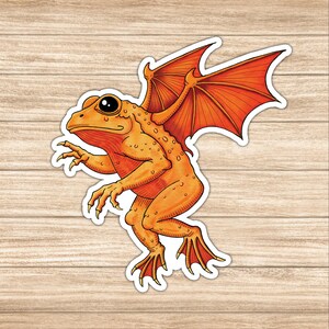 May include: A sticker featuring an orange frog with large, black eyes and orange wings. The frog is in a walking pose with its claws extended. The sticker has a white border and is set against a wood grain background.