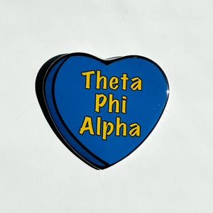 May include: A blue heart-shaped pin with the text "Theta Phi Alpha" in yellow letters.