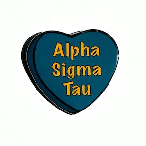 Op de afbeelding: Een tealblauwe hartvormige pin met de tekst "Alpha Sigma Tau" in gele letters.