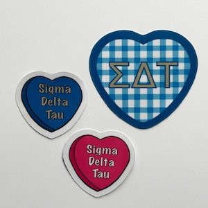 Puede incluir: Tres pegatinas en forma de corazón. Dos son azules con el texto "Sigma Delta Tau". La tercera es rosa con el mismo texto. Una pegatina azul tiene un patrón de cuadros. Las pegatinas están sobre un fondo blanco.