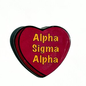 May include: A heart-shaped pin with the words "Alpha Sigma Alpha" in yellow lettering on a maroon background. The pin has a black border and is set against a white background.