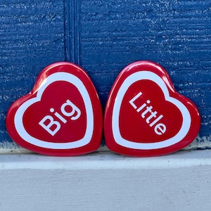 May include: Two red heart-shaped pins with white borders and text. One pin says "Big" and the other says "Little" in white letters. The pins are on a white surface against a blue background.