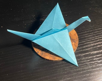 May include: A light blue origami crane perched on a round, brown wooden coaster.