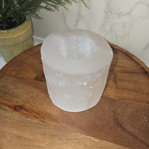 May include: A cylindrical, translucent white selenite candle holder. The candle holder is placed on a round, wooden tray. A small potted plant with green foliage is visible in the background.