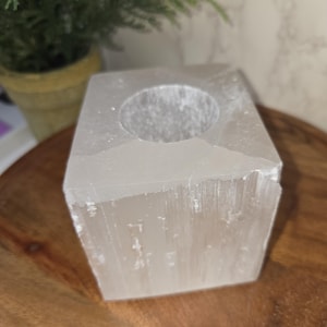 May include: A square, translucent selenite candle holder with a circular opening. The natural stone has a rough, textured surface and sits on a wooden surface. A small potted plant is visible in the background.