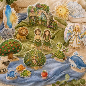 May include: A selection of colourful, illustrated cutouts depicting scenes and characters from a biblical story. Includes Adam and Eve, animals, a tree with apples, a sun, and an angel. The cutouts are arranged on blue and green fabric.