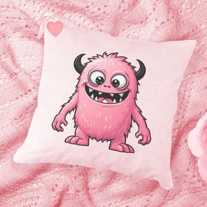 Könnte beinhalten: Ein rosa Kissen mit einem Cartoon-Monster-Design. Das Monster ist rosa mit schwarzen Hörnern, großen Augen und einem breiten Grinsen. Das Kissen ist quadratisch und liegt auf einer rosa Strickdecke.