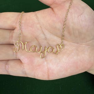 May include: A gold-colored necklace spelling out the name "Maya" in cursive script. The necklace is held in a person's open hand against a dark green background. The chain is delicate and the letters are connected.
