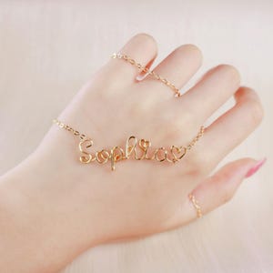 Puede incluir: Un collar de metal en tono dorado con el nombre Sophia escrito en letra cursiva de alambre, acentuado con una pequeña forma de corazón al final. El collar está unido a una delicada cadena dorada que conecta con las letras de alambre.