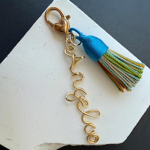 Puede incluir: Un llavero de metal dorado con un cierre de mosquetón en forma de corazón y un nombre formado con alambre que dice "Angela". Unido al cierre hay una borla de tela multicolor con hilos en azul brillante, verde oliva, beige y rojo.