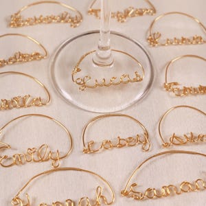 May include: A collection of gold-coloured wire wine glass charms, each shaped into a unique name in cursive script. One charm is attached to the base of a clear glass stem, while others are arranged around it, featuring names like Lucas, Emily, and Samantha.
