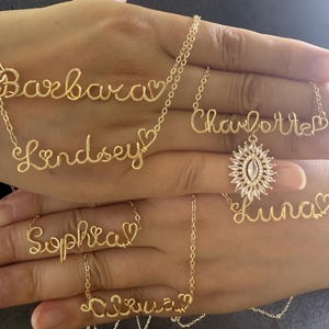 Puede incluir: Una colección de collares con nombres en alambre de tono dorado que muestran los nombres Barbara, Lindsey, Charlotte, Luna, Sophia y Olivia. Cada nombre está en cursiva con un pequeño corazón y un collar incluye un colgante de sol.
