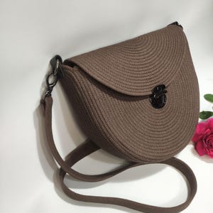 May include: A brown, semi-circular handbag with a woven texture. The bag features a flap closure with a black clasp and a matching brown shoulder strap. The bag is designed with a rounded shape and a simple, elegant style.