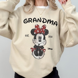 Op de afbeelding: Beige sweatshirt met het woord "GRANDMA" in zwart, met een Minnie Mouse-afbeelding met een rode stippen strik. De tekst "est. 2026" en het Disney-logo zijn ook zichtbaar.