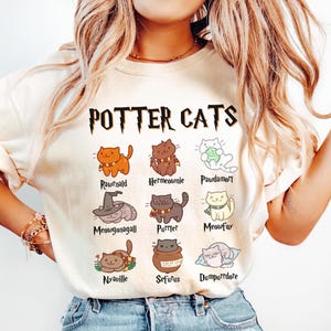 Puede incluir: Una camiseta blanca con el texto "POTTER CATS" en letras negras y amarillas. La camiseta presenta una cuadrícula de nueve gatos de dibujos animados con nombres como "Rawnold", "Hermeownie" y "Meowfoy".