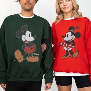 May include: Two sweatshirts featuring Mickey and Minnie Mouse. One is dark green with Mickey Mouse in red shorts and brown shoes. The other is red with Minnie Mouse in a polka dot skirt and brown shoes. Both have the Disney logo.